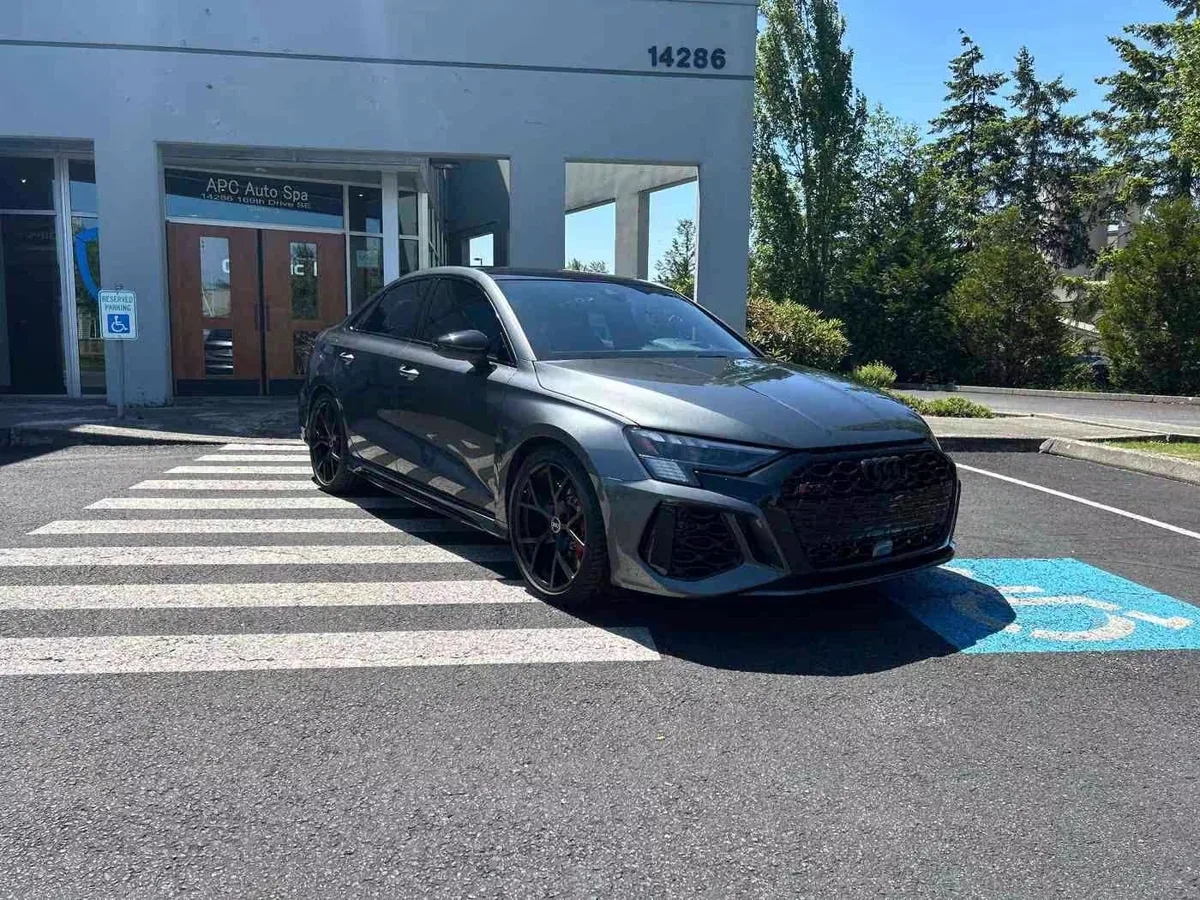 A gray Audi sedan parked outside the APC Auto Spa shop entrance on a sunny day